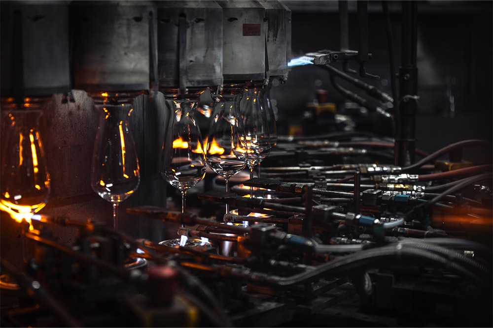 Reihe von Gläsern in einer industriellen Glasproduktionsanlage, umgeben von Metallarmen und glühenden Lichtreflexen während des automatisierten Fertigungsprozesses.