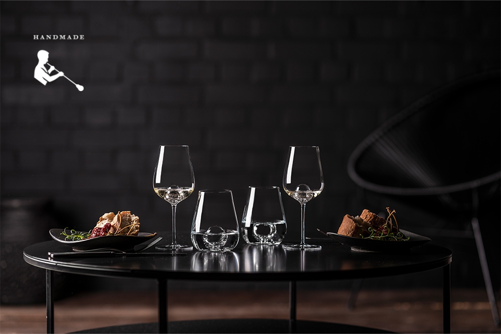 Handmade glasses from the Air Sense series by Zwiesel Glas, filled with white wine and water, placed on a table with a dark background; small dishes on black plates enhance the elegant setting.