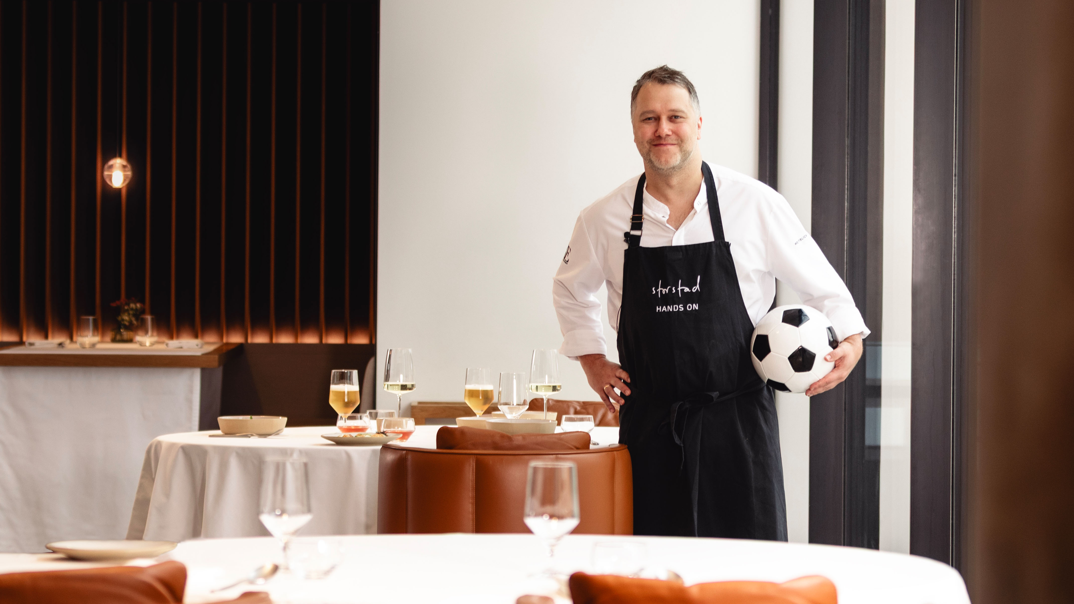 Ein Koch, der einen Fußball unter dem Arm hält, steht in einem stilvoll eingerichteten Restaurant mit elegant gedeckten Tischen.