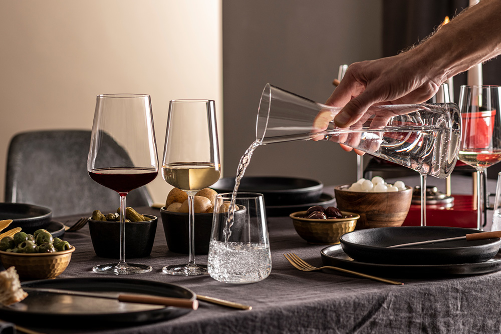 Red wine, white wine and drinking glasses from the Vervino range on a table laid with food. Water is being poured into the water glass.