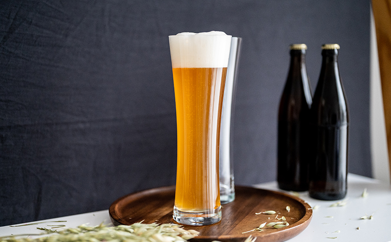 Un verre de bière mousseux est posé sur un plateau en bois recouvert de brins de blé.