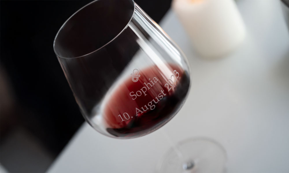 Un verre à vin rouge gravé « Sophia 10 août 2026 » est posé sur une table claire à côté d'une bougie.