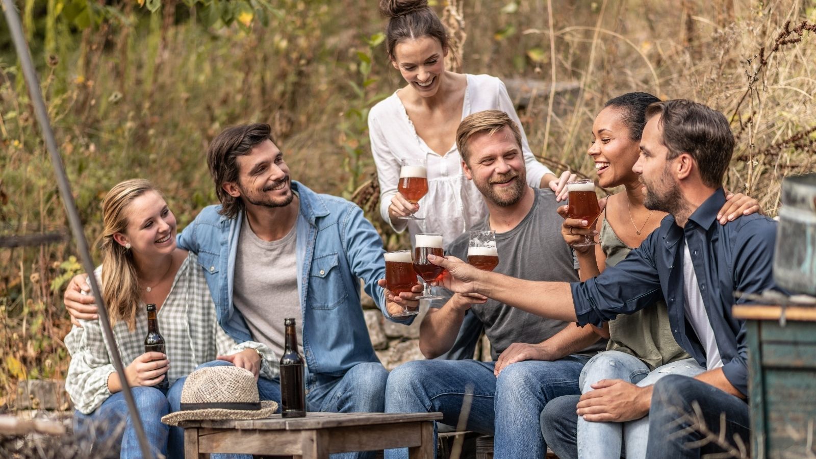 Six people clink beer glasses while sitting together outdoors at a wooden table.