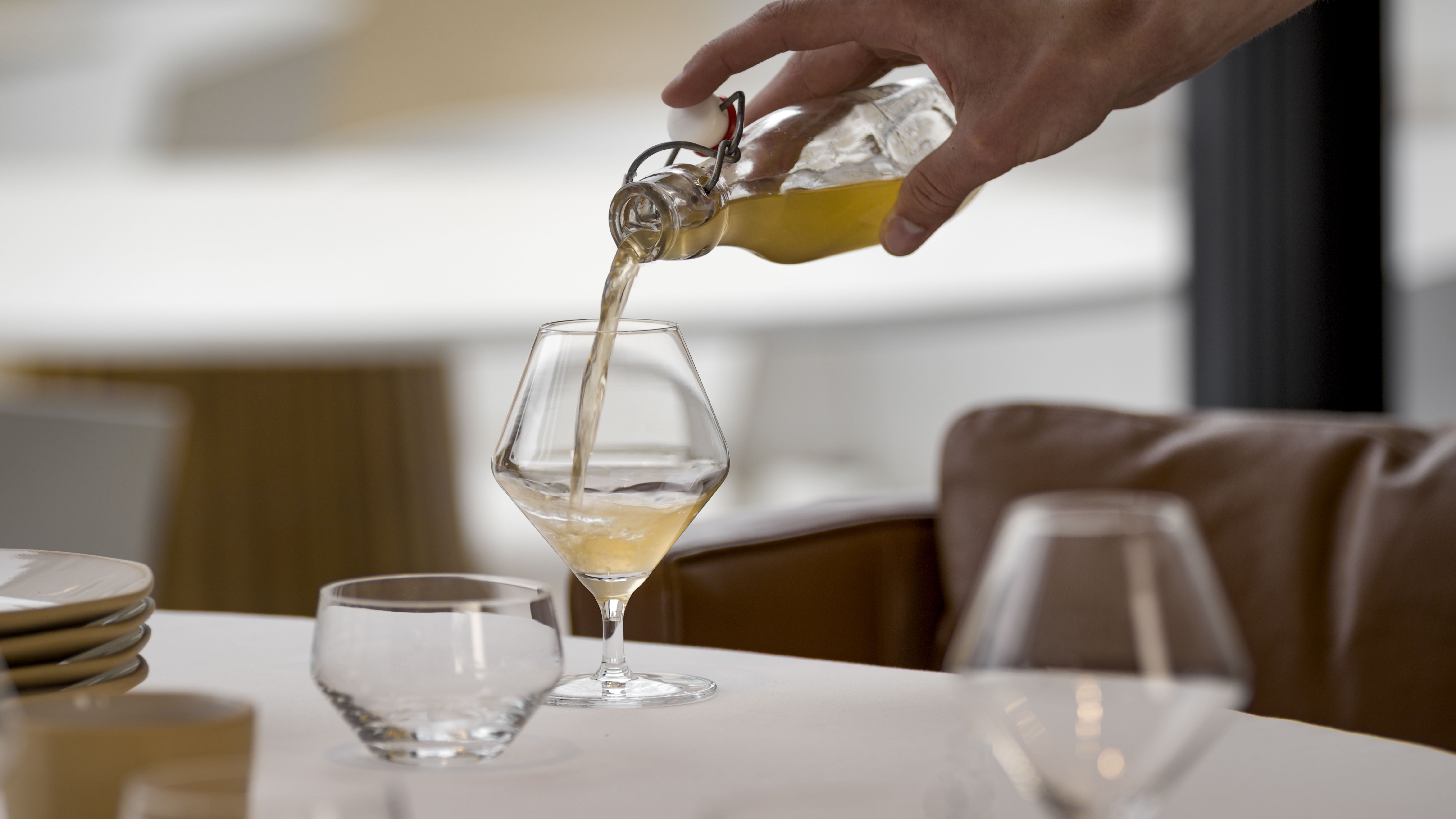 Ein Getränk wird in ein Glas eingeschenkt, daneben stehen weitere Gläser auf einem gedeckten Tisch.