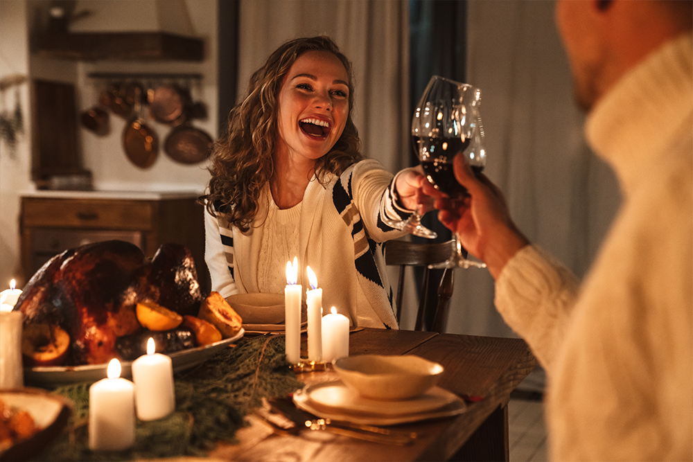 Vrouw proost met man aan feestelijk gedekte tafel met kaarsen en kalkoen, beide lachen.