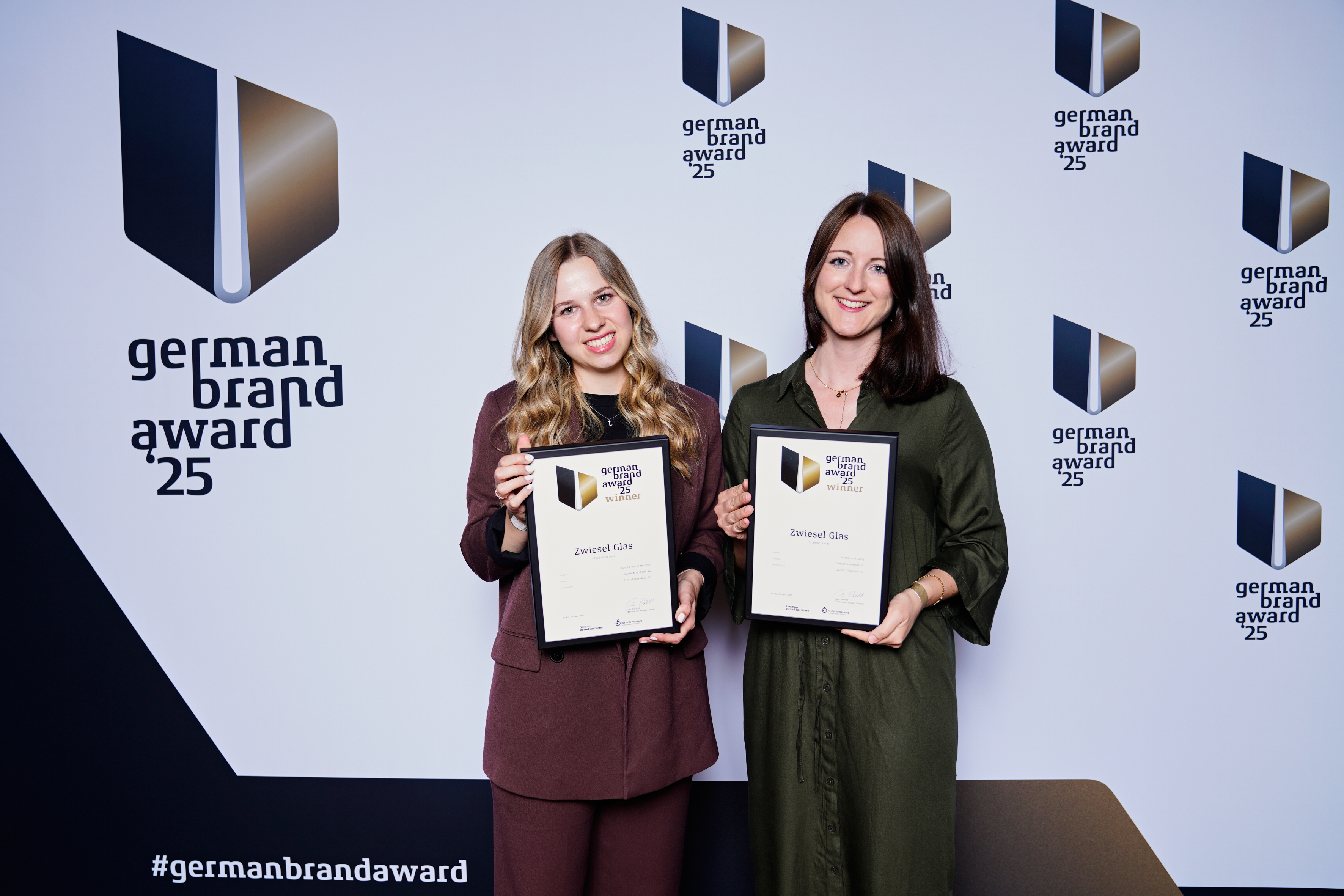 Eva Wittenzellner und Lisa Müller halten Urkunden des German Brand Award 2025 vor einer Wand mit Logo und Schriftzug des Awards.