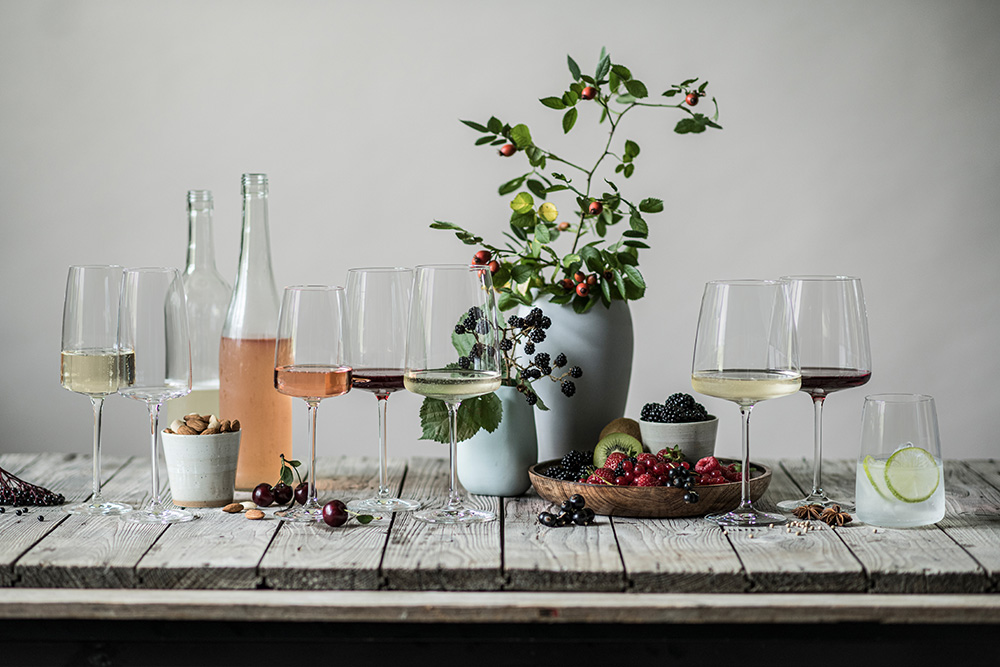 All the glasses from the Vivid Senses range on a wooden table. Filled with various wines. A wine vase with decorative branches in the background.