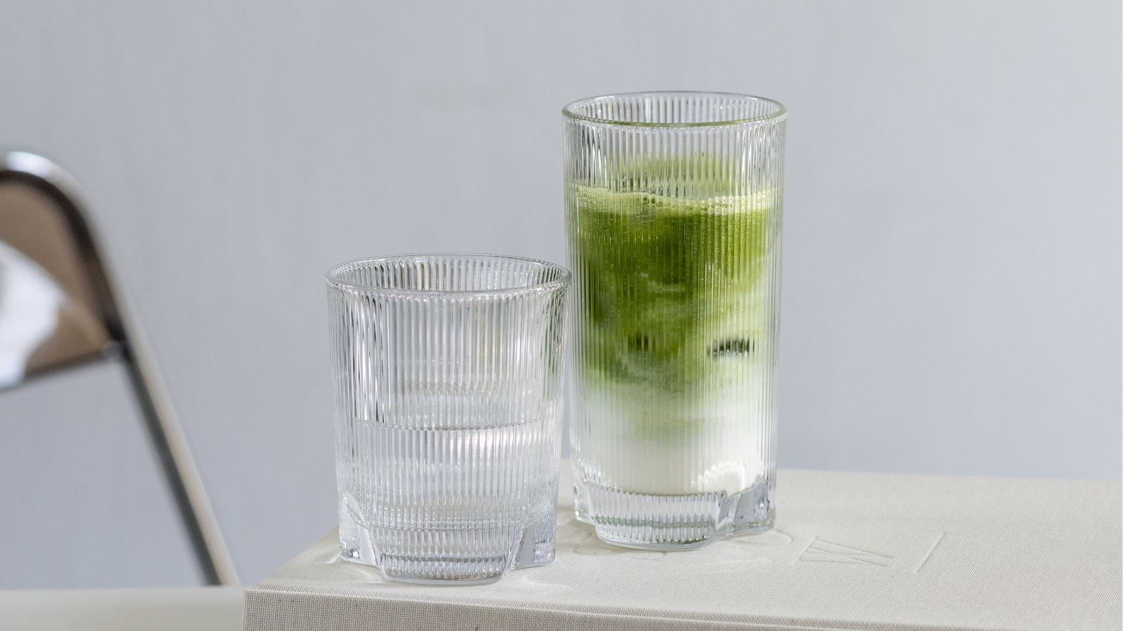 Two ribbed glasses on a light-colored table, one filled with an Iced Matcha Latte.
