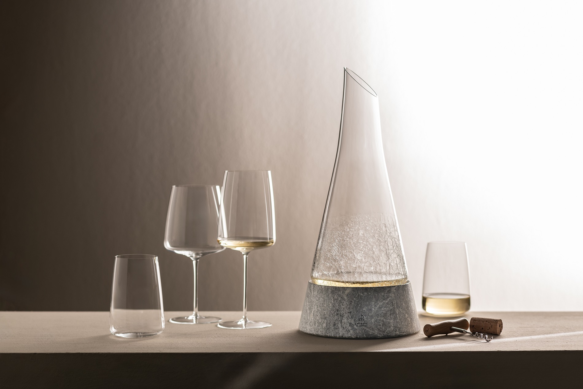 Elegante Weingläser und Dekanter in Steinsockel mit Weißwein auf einem Tisch, daneben Korkenzieher und Korken vor neutralem Hintergrund.