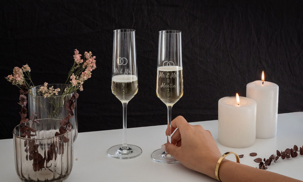 Two engraved sparkling wine glasses stand next to flowers and burning candles on a festively arranged white table.