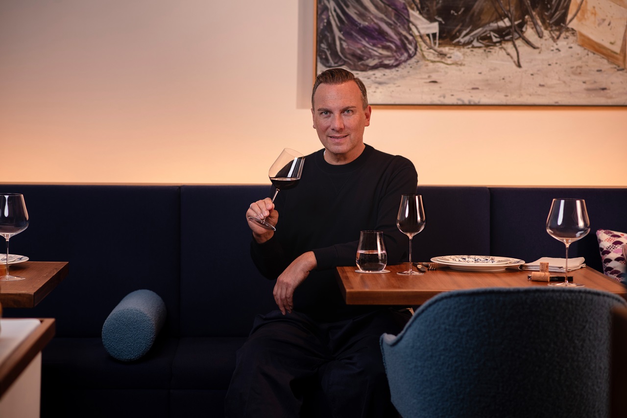 Tim Raue sitzt an einem gedeckten Tisch in modernem Restaurantambiente und hält ein Glas Rotwein in der Hand.