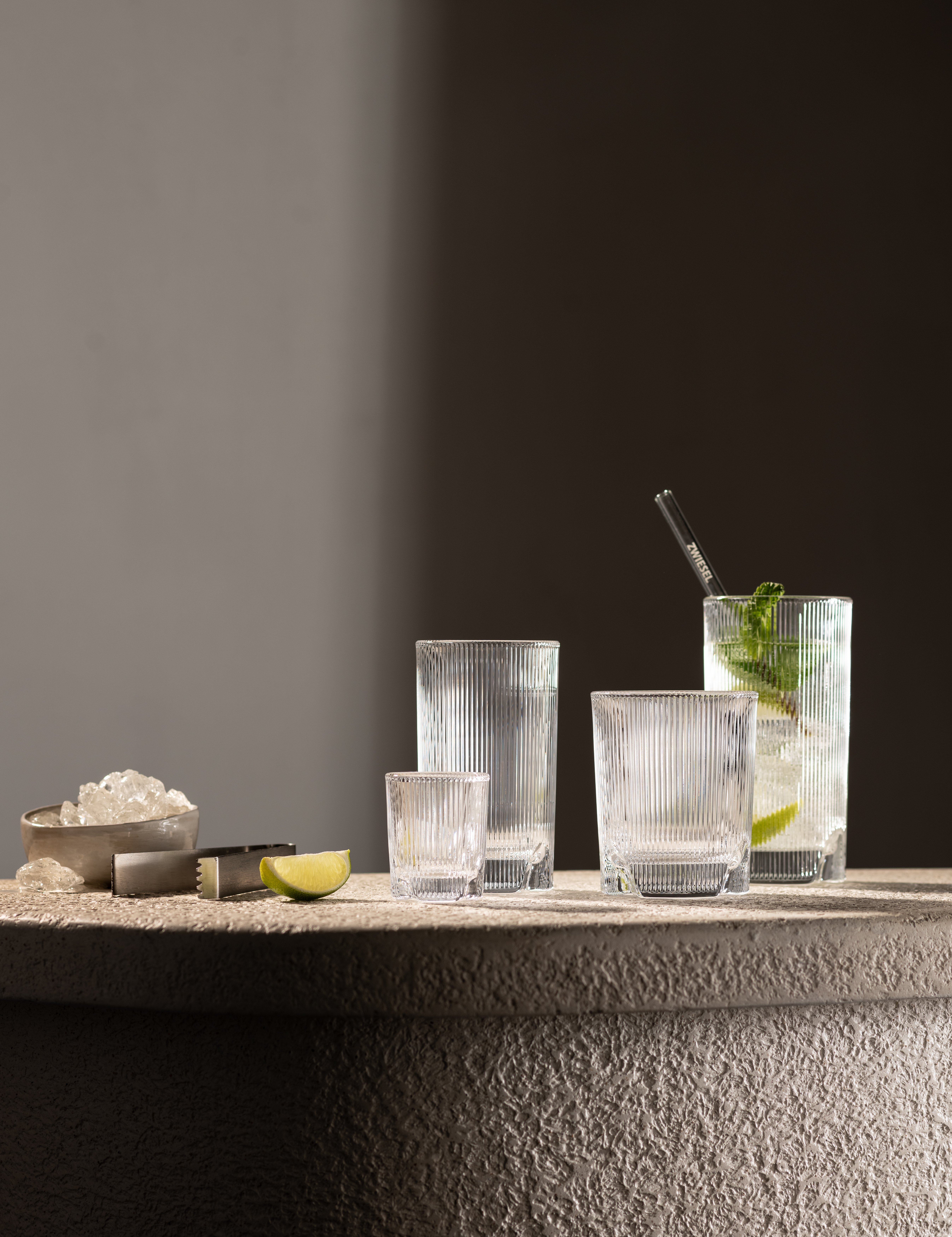 Vier Gläser der Serie Fave von Schott Zwiesel auf heller Steinplatte, mit Limettenstück, Eiswürfeln und Eiszange, im warmen Licht.