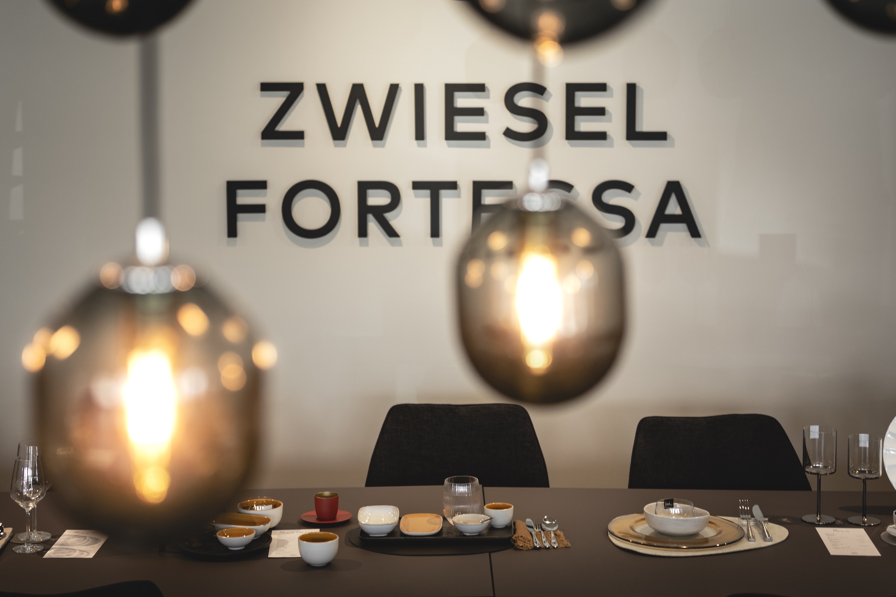 Gedeckter Tisch und ZWIESEL FORTESSA-Logo im Hintergrund. Im Vordergrund zwei Glaslampen, die von der Decke hängen. Gemütliche Atmosphäre.