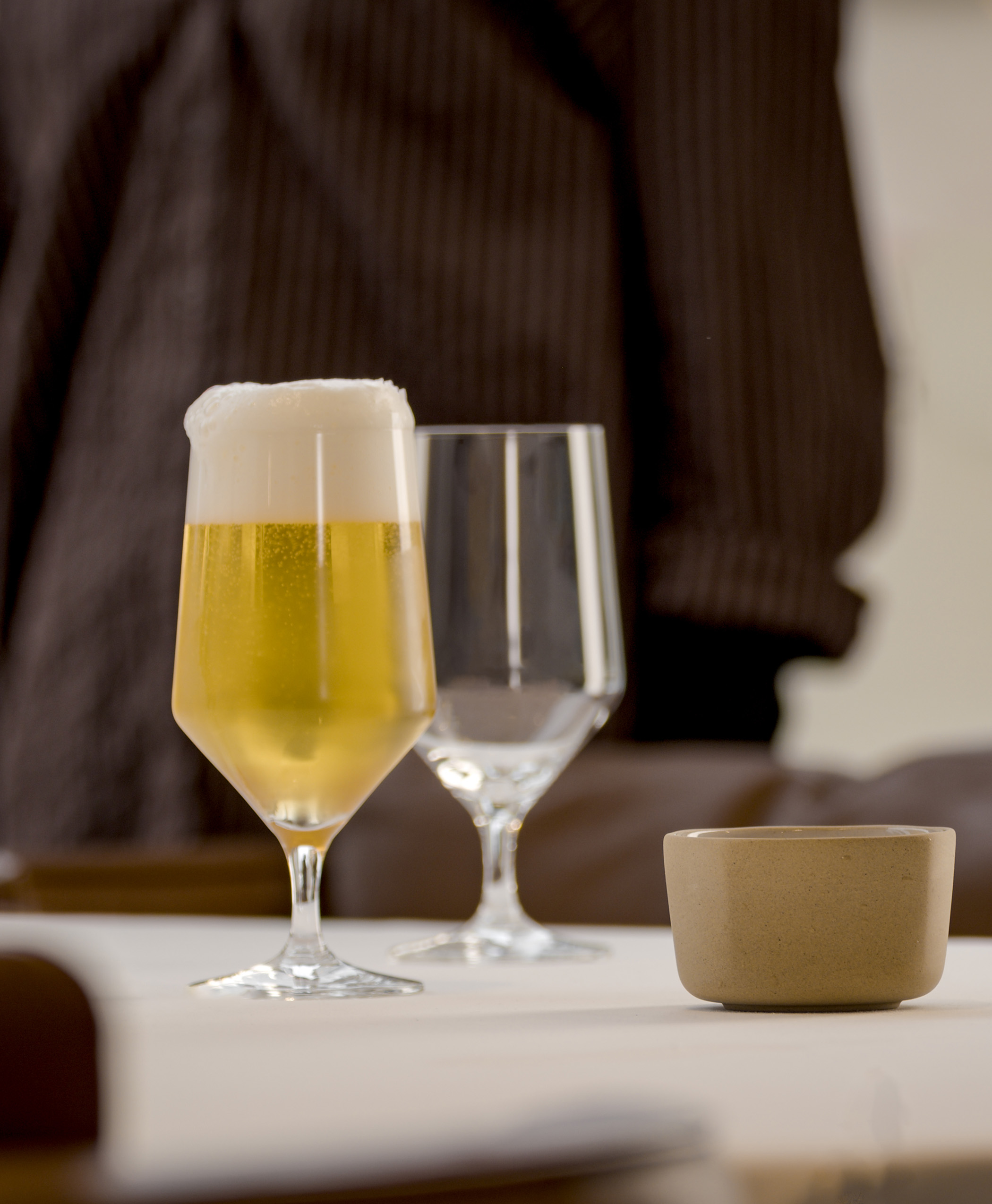 Glas mit hellem Bier und Schaumkrone auf einem gedeckten Tisch, daneben ein weiteres Glas und eine Bowl.