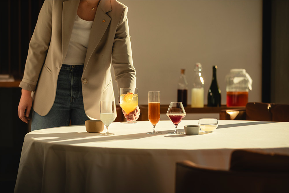 Eine Person stellt ein Glas mit orangem alkoholfreiem Getränk auf einen weiß gedeckten Tisch, auf dem bereits mehrere unterschiedlich geformte Gläser mit farbigen Drinks stehen.