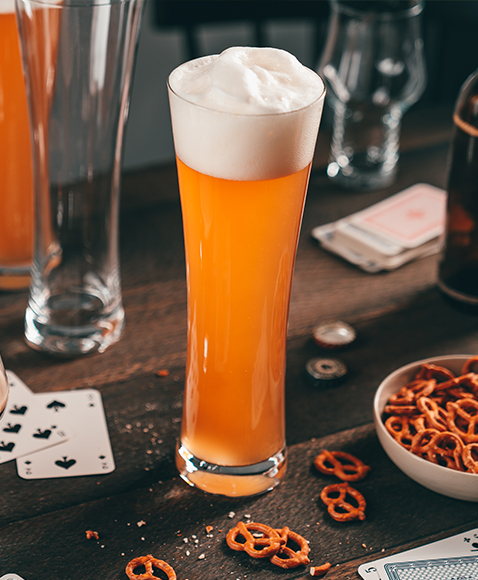 Wheat beer glass with beer and foam head on a rustic wooden table, plus pretzels, playing cards, and more beer glasses.