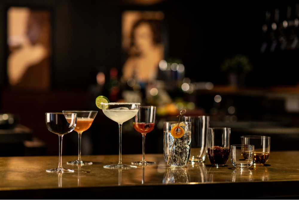 Various elegant glasses from the Tone bar series by Zwiesel Glas are arranged decoratively on a bar with colorful drinks.