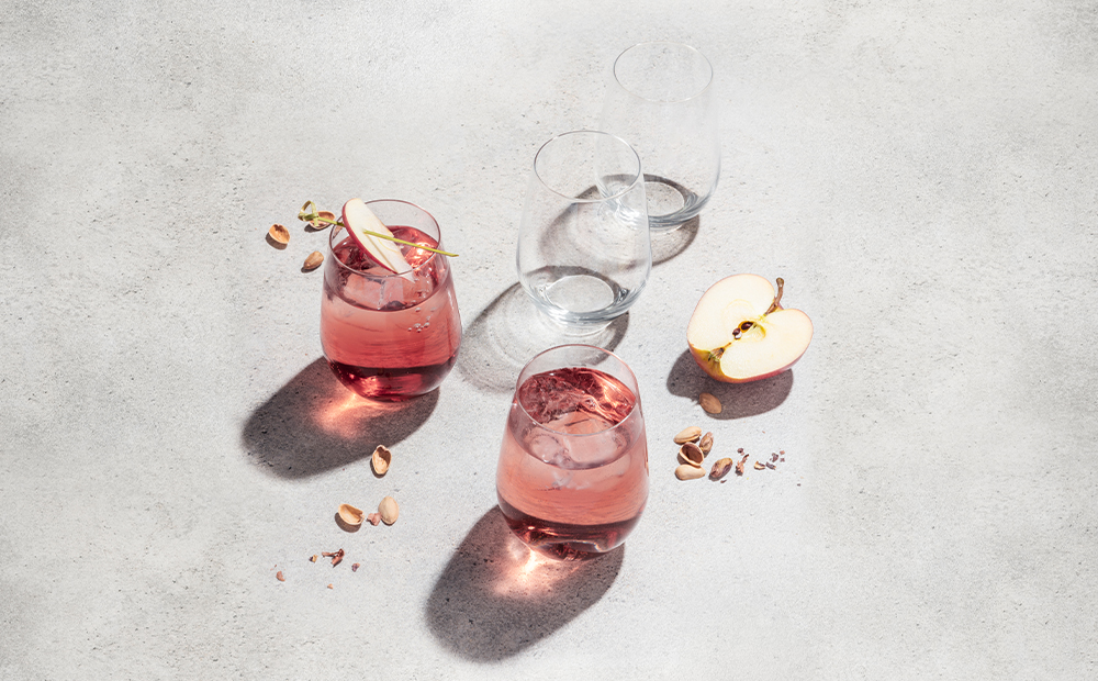 Zwei Gläser mit rosa Getränk, Eis und Apfelscheiben, leere Gläser, Pistazien und Apfelhälfte auf hellem Tisch, Sonnenlicht.