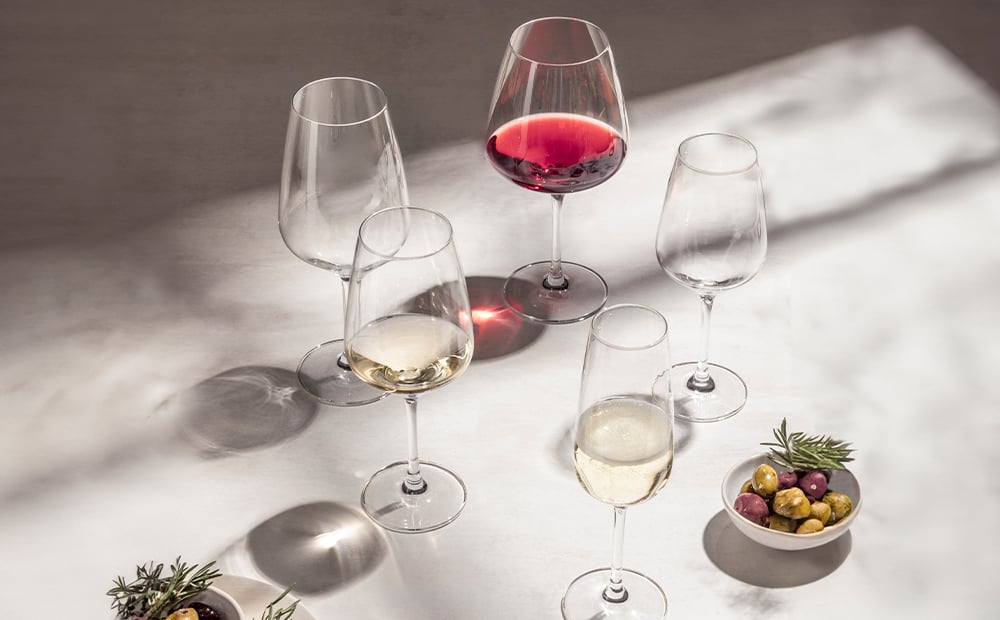 Gläser mit Rotwein, Weißwein und Sekt, Oliven in Schale und Rosmarin auf hellem Tisch, Sonnenlicht wirft Schatten.