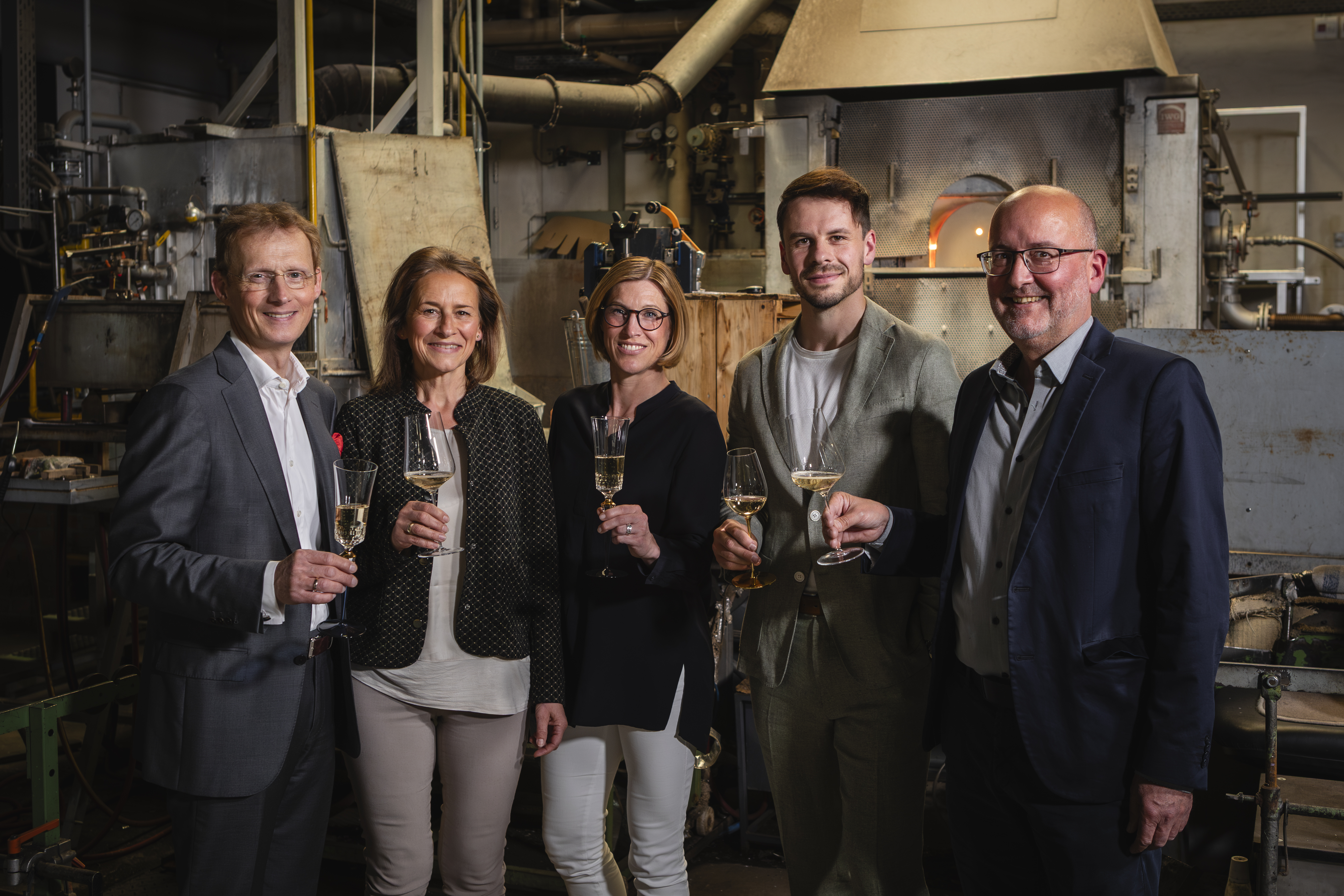 Fünf Personen stehen in einer Glasmanufaktur und halten Sektgläser, umgeben von industriellen Maschinen und warmem Licht.