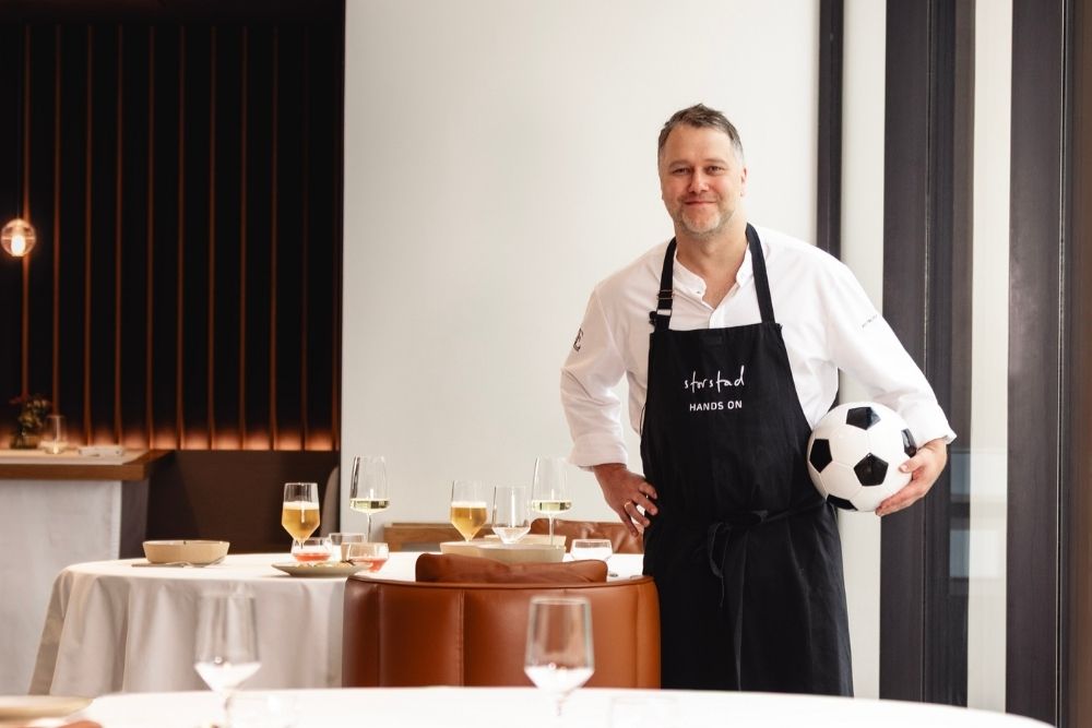 Ein Koch, der einen Fußball unter dem Arm hält, steht in einem stilvoll eingerichteten Restaurant mit elegant gedeckten Tischen.