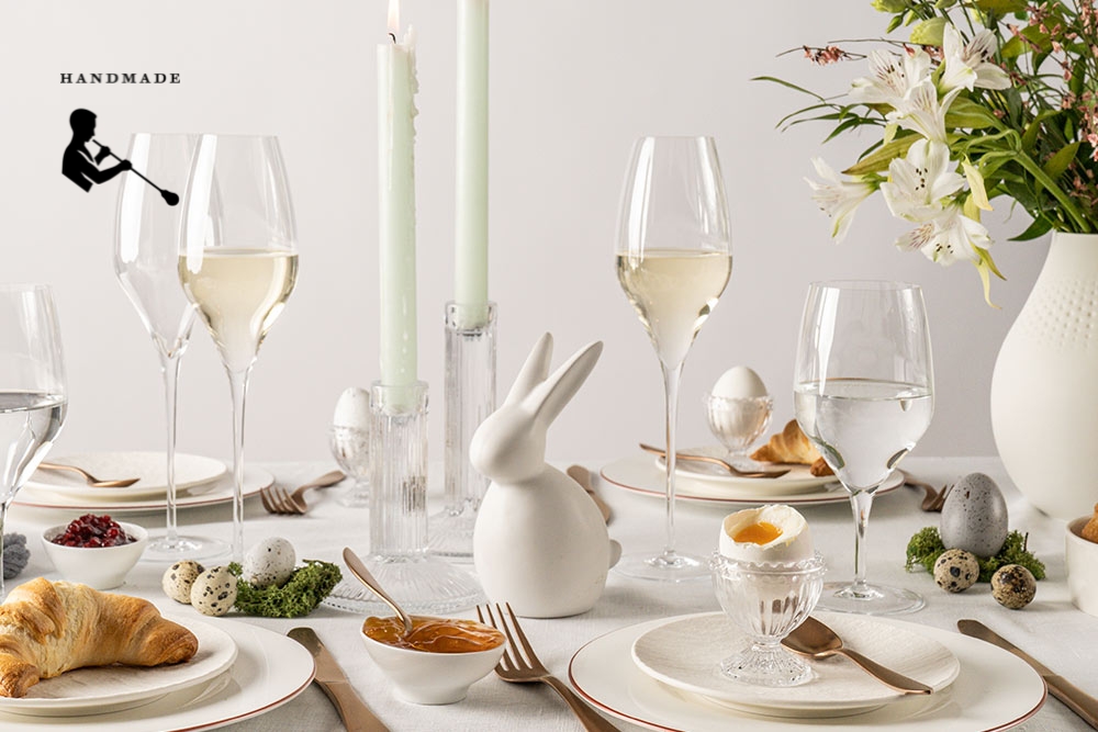 Handmade glasses from the Alloro series by Zwiesel Glas, filled with champagne/sparkling wine and water, on a festively decorated Easter breakfast table with a white tablecloth, Easter decorations, croissants, flowers, and candles.