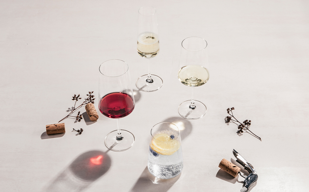 Gläser mit Rotwein, Weißwein und Wasser, Korken, Korkenzieher und Zweige auf hellem Tisch, Sonnenlicht wirft Schatten.