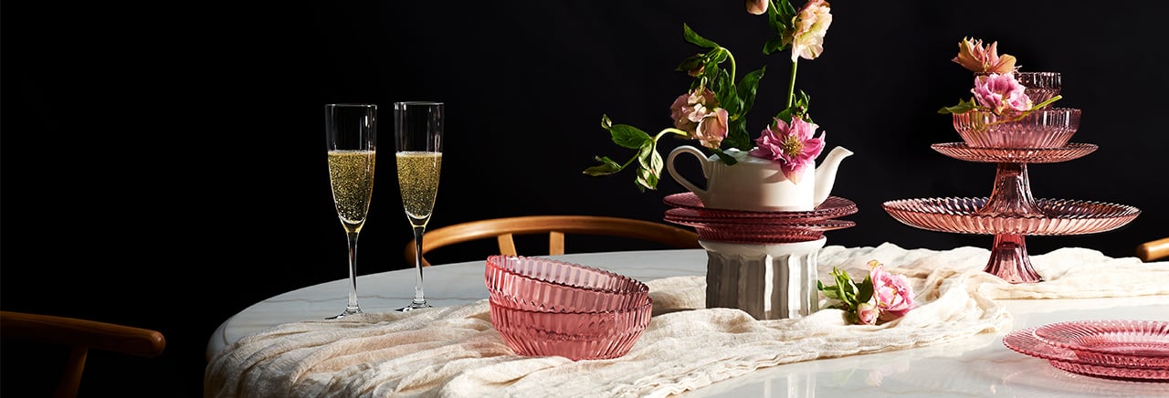 Tafel versierd met bloemen, twee champagneglazen en roze glazen servies. Het servies bestaat uit dessertschaaltjes, borden en een etagère