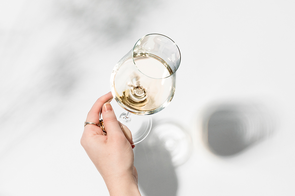 Main tenant un verre à vin blanc Duo de Zwiesel Glas rempli de vin blanc au-dessus d’une surface blanche, lumière et ombre visibles.