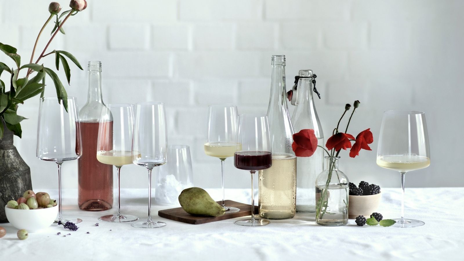 Elegante Weiß- und Rotweingläser in verschiedenen Glasgrößen und Formen, stilvoll arrangiert auf einem gedeckten Tisch mit Flaschen, Früchten und floralen Akzenten.