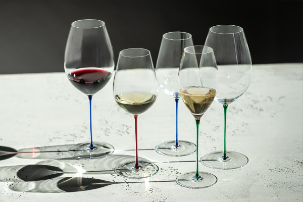 Verres à pied de la série artisanale Enoteca Colors de Zwiesel Glas avec des pieds colorés en vert béryl, rouge cerise et bleu clair, disposés sur une table lumineuse et remplis de vin rouge, vin blanc et champagne.