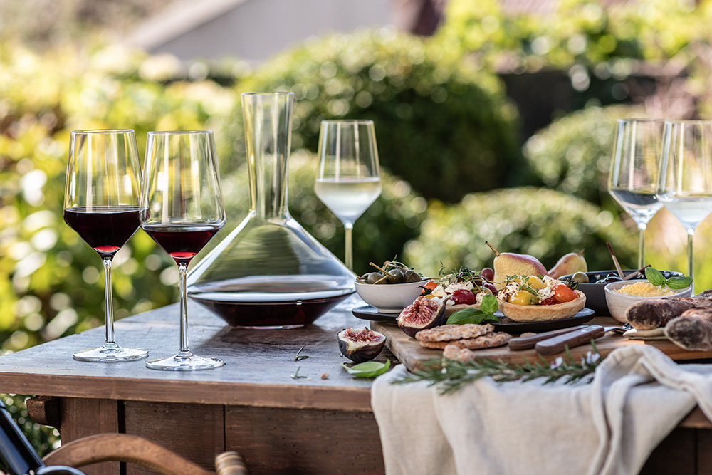 Red wine and white wine glasses, and the decanter from the Pure range, on a wooden table in the garden with food.