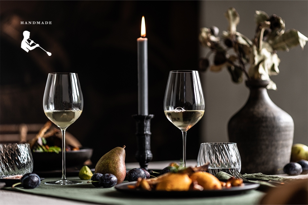 Wine glasses from the handmade Enoteca series by Zwiesel Glas, filled with white wine, and tumblers from the Manoa series, filled with water, stand on a table with autumnal dishes, a burning candle, and vases in the background.