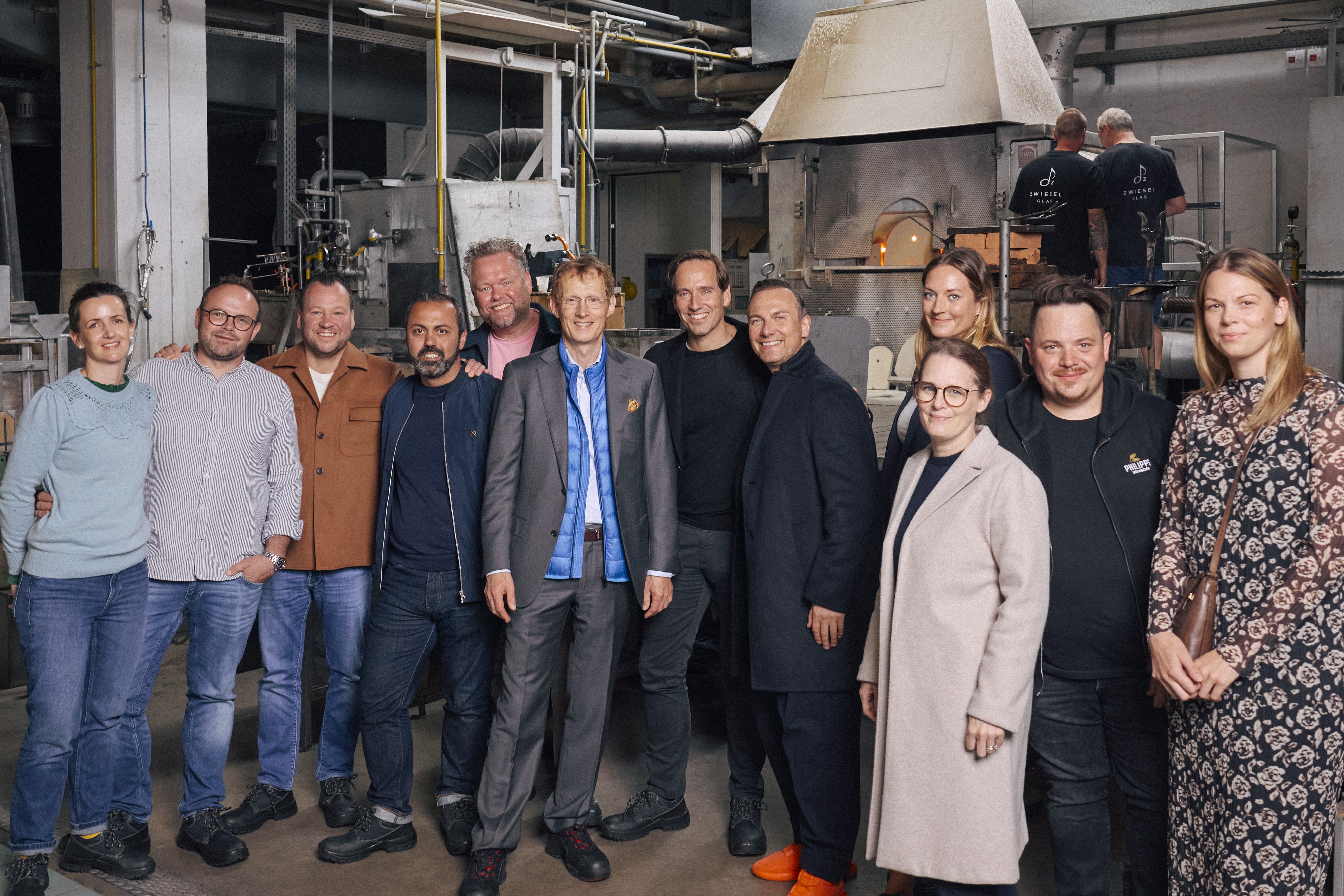 Eine Gruppe von Personen steht in einer Glasmanufaktur vor einem Ofen, umgeben von Maschinen und industriellem Ambiente.