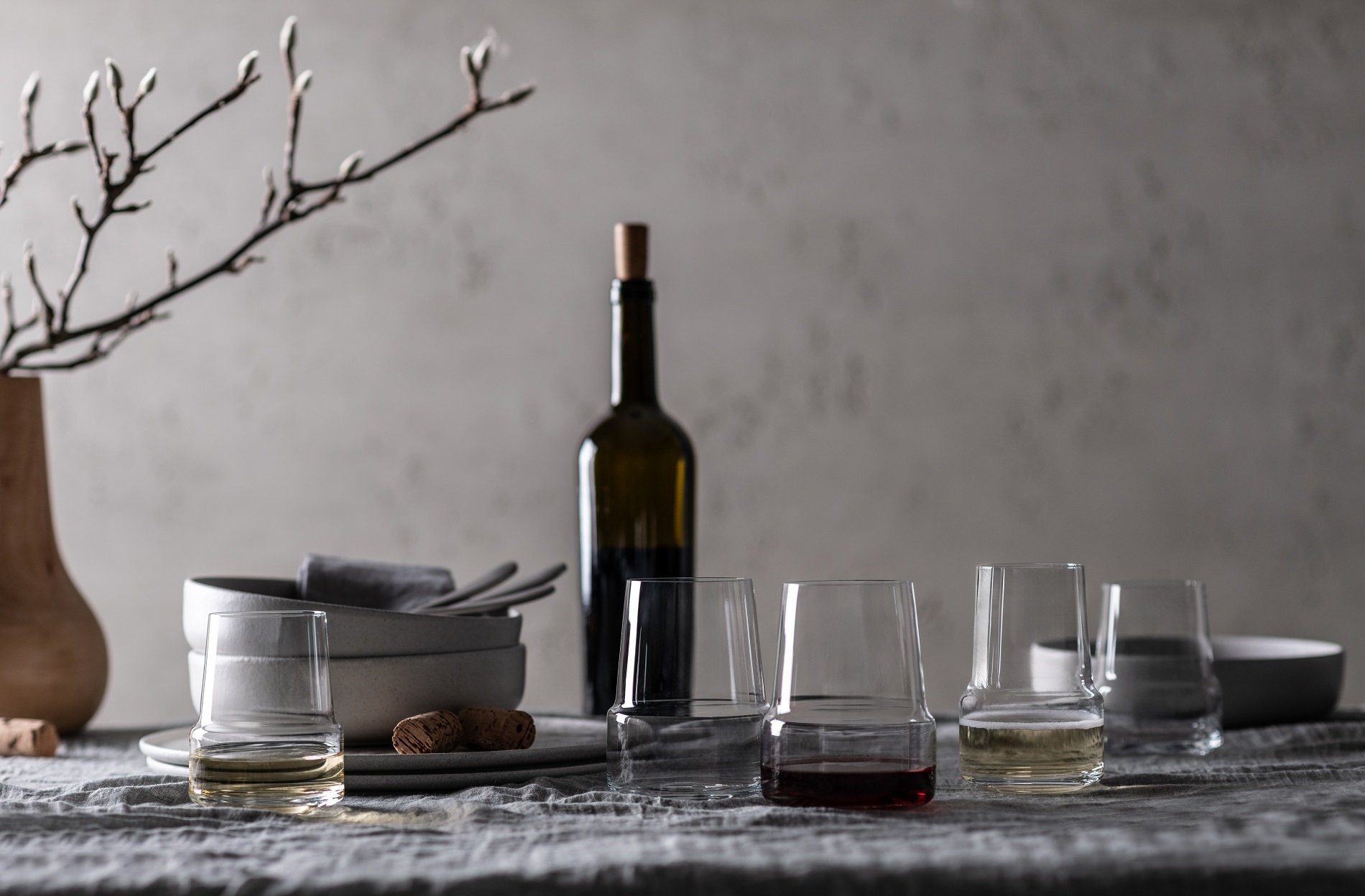 Eleganter Tisch mit Wein-Tumblern, Weinflasche, Schalen und Zweigen in Vase vor grauer Wand.