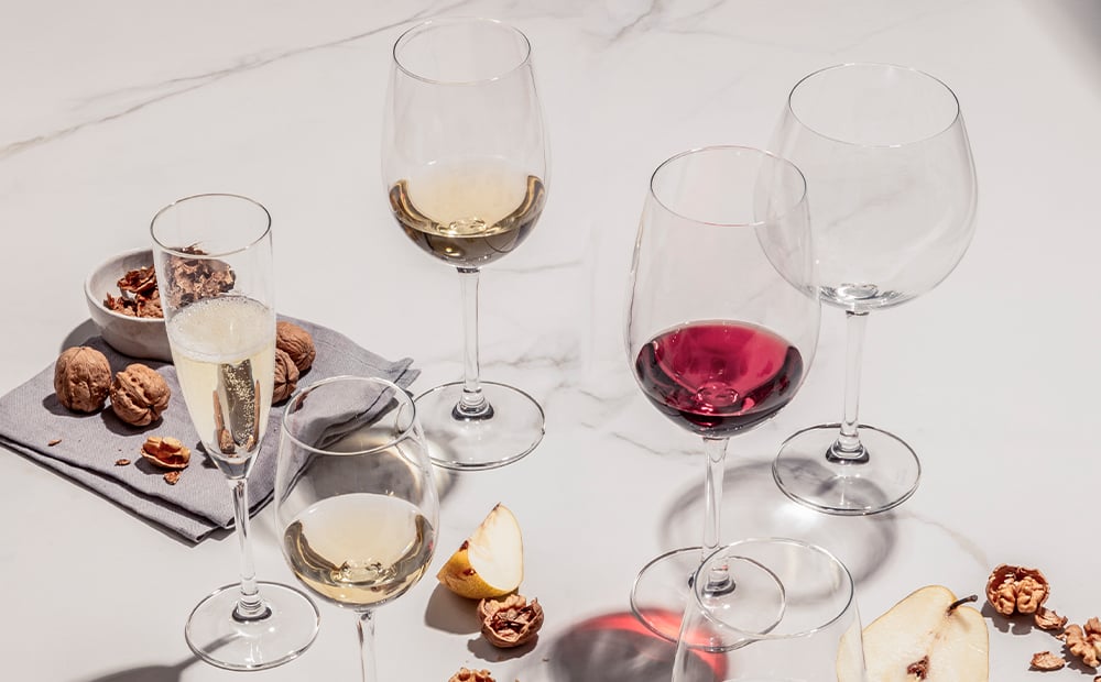 Weingläser mit Rot- und Weißwein, Sekt, Walnüsse und Birnenscheiben auf hellem Tisch, Sonnenlicht wirft Schatten.