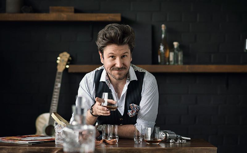 Sommelier Justin Leone at a bar with handmade Enoteca bar glasses by Zwiesel Glas, filled with whisky; relaxed, creative atmosphere with musical instruments in the background.
