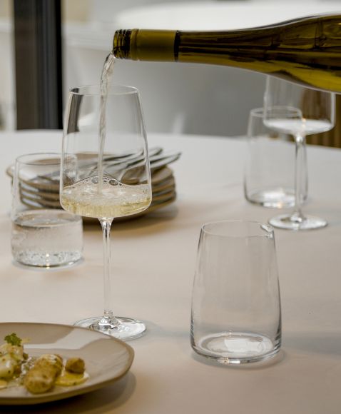 Weißwein wird in ein Weinglas eingeschenkt, umgeben von weiteren Gläsern und Geschirr auf einem gedeckten Tisch.