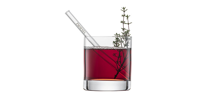 Un verre contenant une boisson rouge, une paille en verre et une branche d'herbe aromatique.