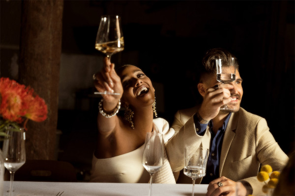 Deux personnes souriantes lèvent leur verre pour trinquer, l'ambiance est festive avec des fleurs en arrière-plan.