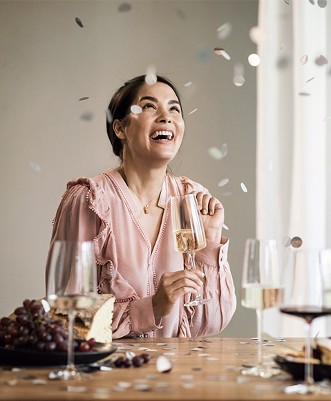 Smiling woman in a pink blouse at the table, holding a sparkling wine glass from the Simplify series by Zwiesel Glas; confetti, cake, and grapes around her.