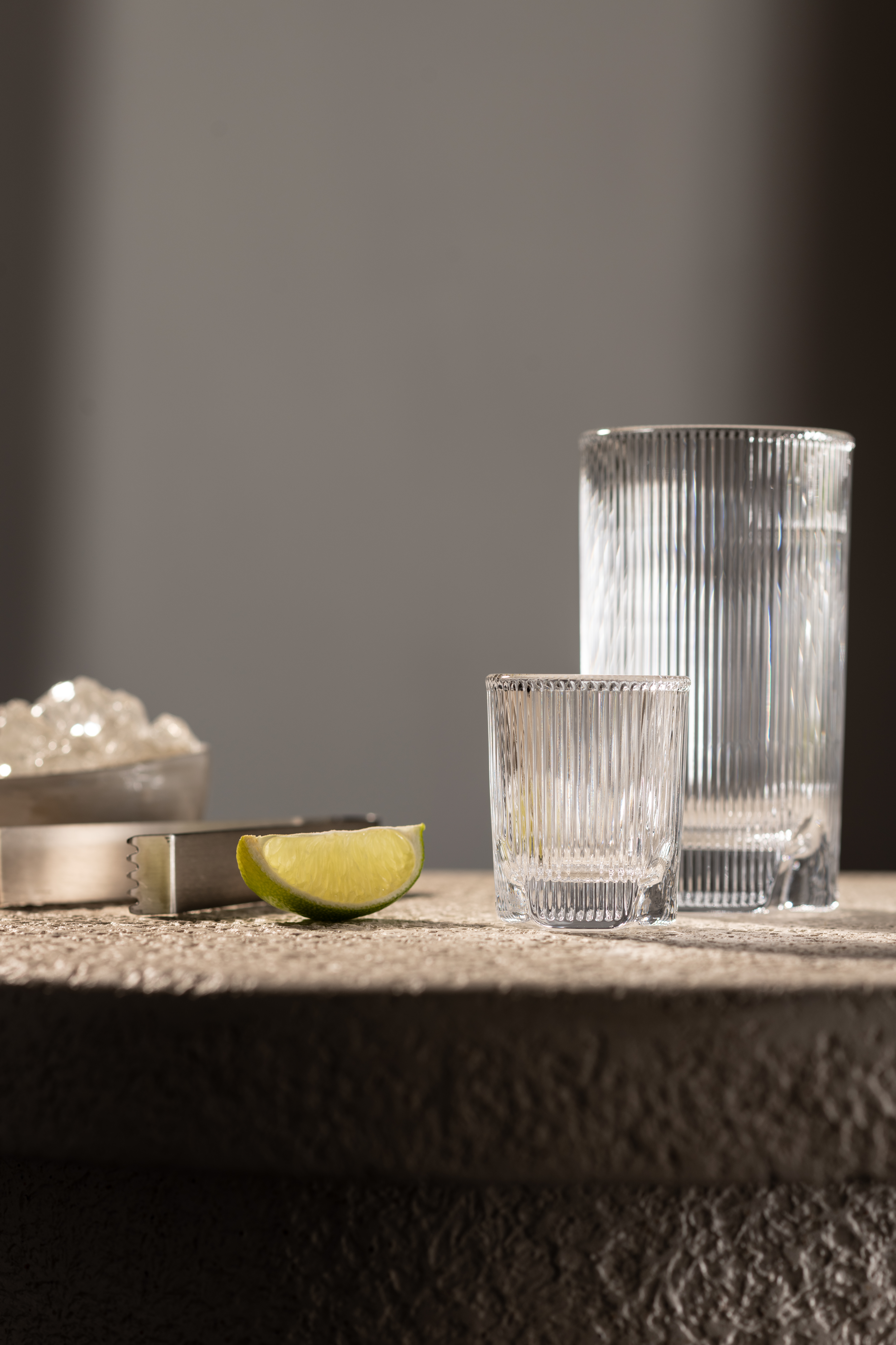 Gläser der Serie Fave von Schott Zwiesel auf heller Steinplatte, mit Limettenstück, Eiswürfeln und Eiszange, im warmen Licht.
