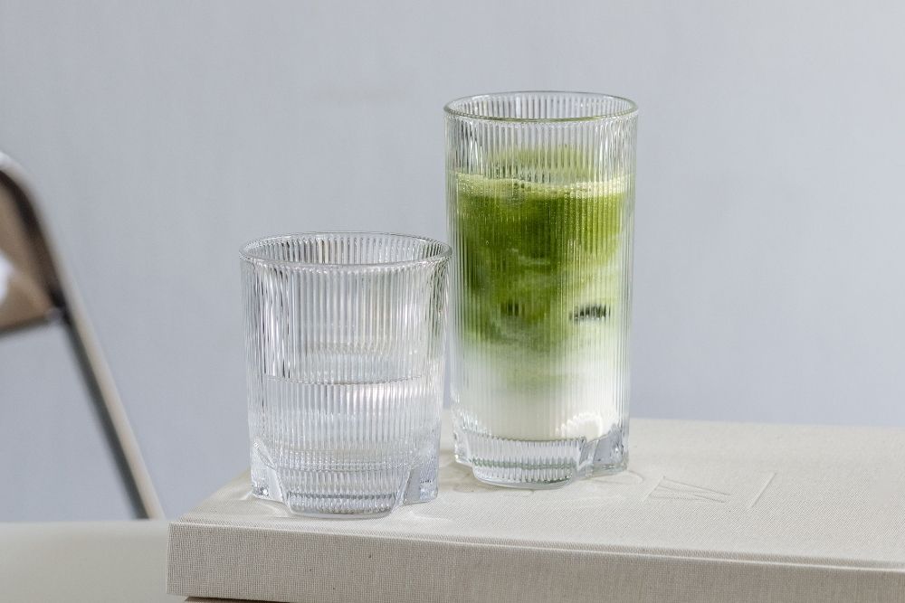 Zwei geriffelte Gläser auf einem hellen Tisch, eines gefüllt mit Iced Matcha Latte.